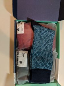 The opened shipping box has three items inside: a pair of blue and green socks with a design on them and two lounge shorts in plastic bags, one red and the other black.