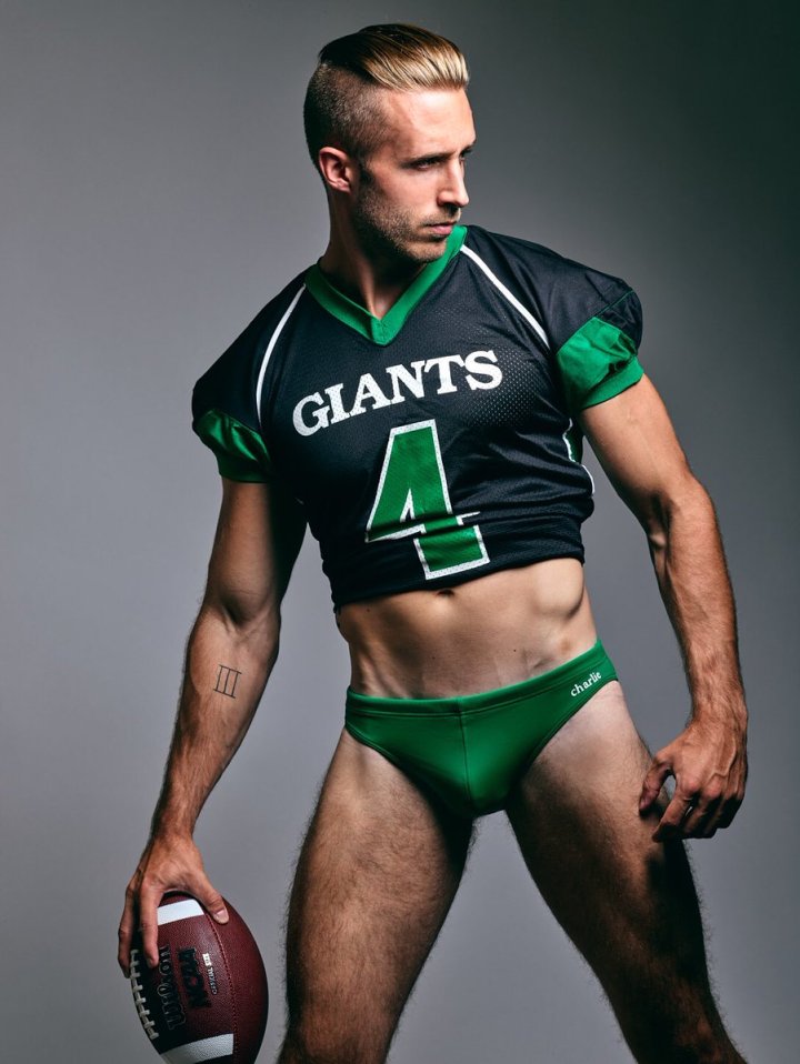 Model, actor, and Texas-based photographer Dillion Lalor posed in a green and black American football jersey and holds a football in his left hand while looking at something on the right side of the image. Dillion wears green swim briefs from the charlie brand.